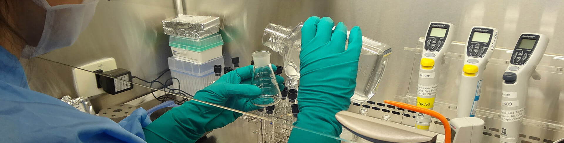 Imagem em close nas mãos de um cientista com luvas verde-água tranferindo líquidos entre recipientes
