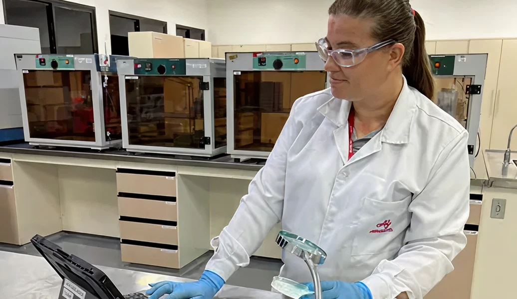 Mulher com jaleco branco e luvas azuis, dentro de um laborátorio