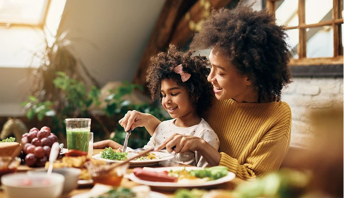 Mãe comendo salada com sua filha em frente a uma mesa repleta de comidas