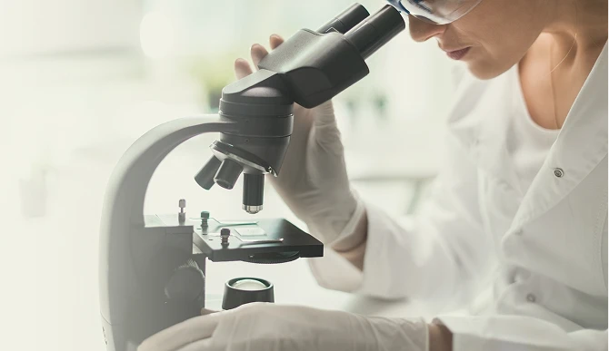 Uma cientista de jaleco branco e óculos de proteção operando um microscópio de alta precisão para analisar minuciosamente uma lâmina de laboratório.