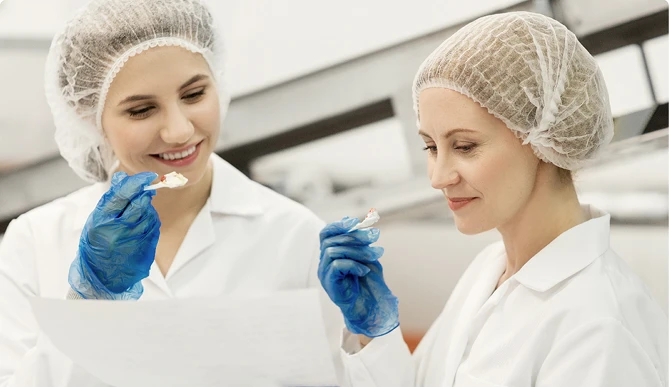 Duas cientistas em um laboratório, usando jalecos, toucas e luvas azuis, examinando documentos e pequenas amostras enquanto sorriem, sugerindo um processo de controle de qualidade ou degustação técnica.